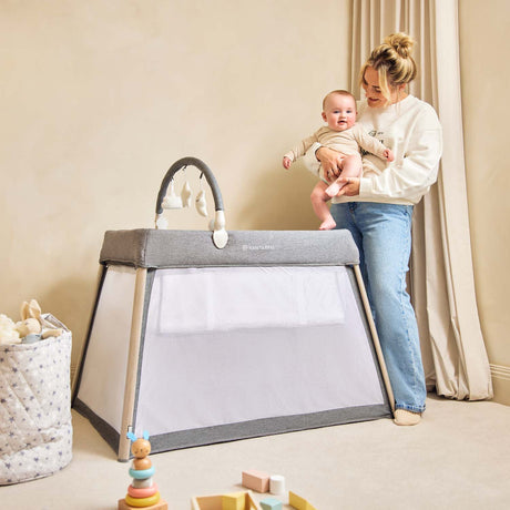 Mom carrying her baby while standing beside the Ickle Bubba Scout 3-in-1 Travel Crib, Cot and Playpen