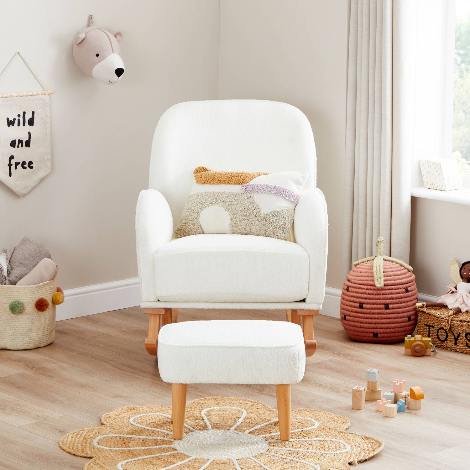 A straight-on front shot of Babymore Freya Nursing Chair with Footstool Set in Off White Boucle colour in a corner of a nursery room with a basket full of baby linens and wooden toys on the floor.