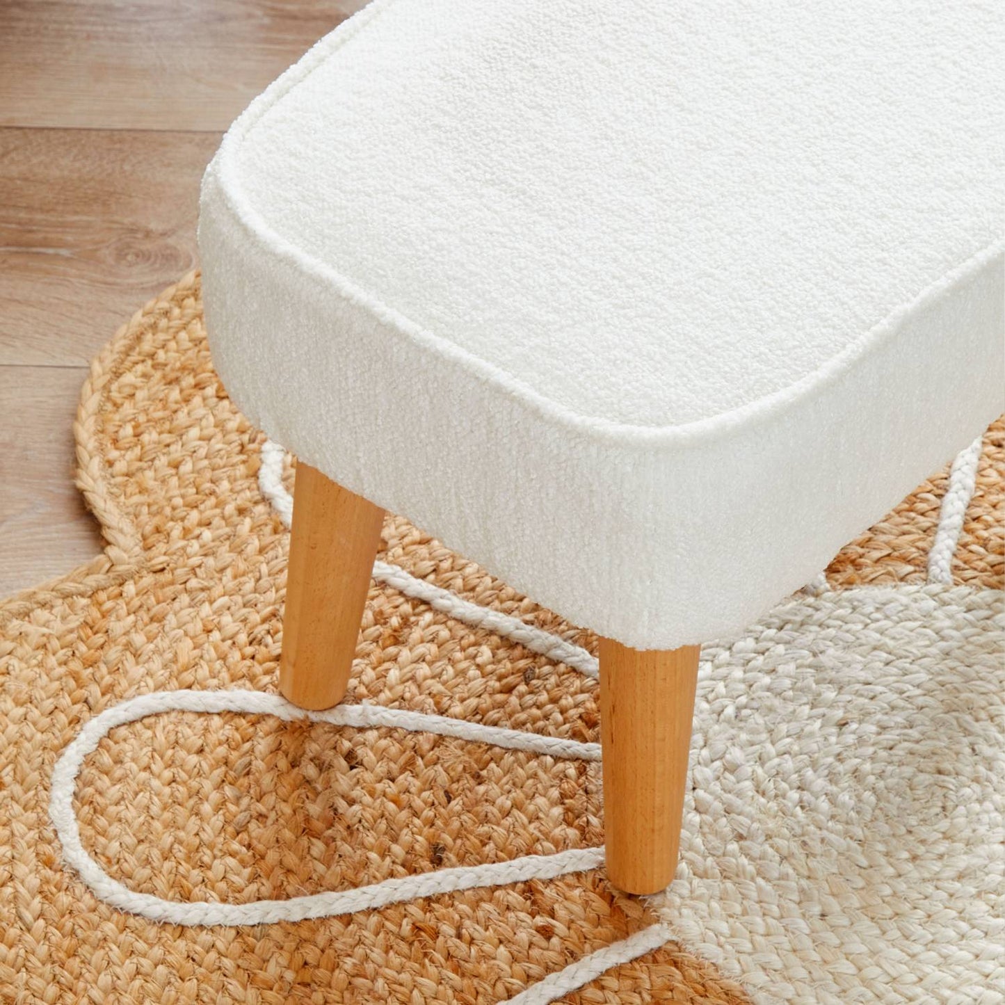 A close-up shot of the Babymore Freya Footstool in Off White Boucle color placed on a natural fiber rug and showing the natural solid beech wood legs.