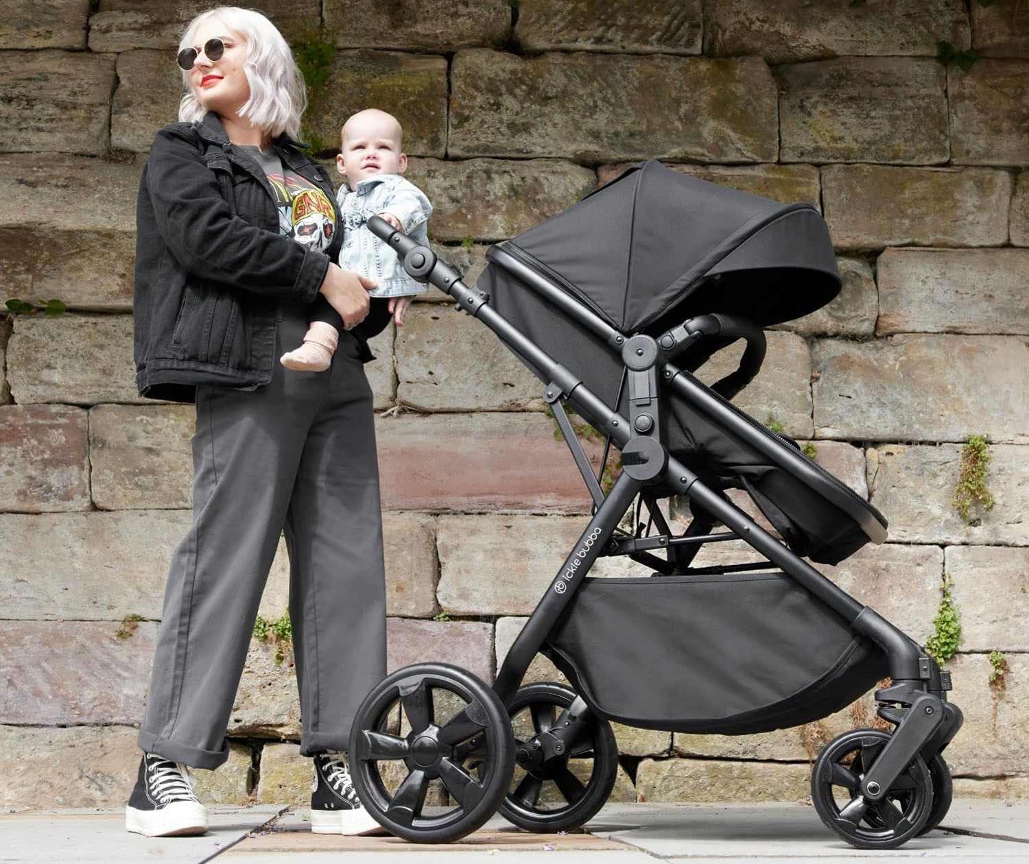 A mother holding her baby beside an Ickle Bubba Comet 3-in-1 Travel System pushchair in Black colour