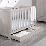 Angled lifestyle shot of Ickle Bubba Pembrey Cot Bed with opened Under Drawer storage full of clothes in Ash Grey colour