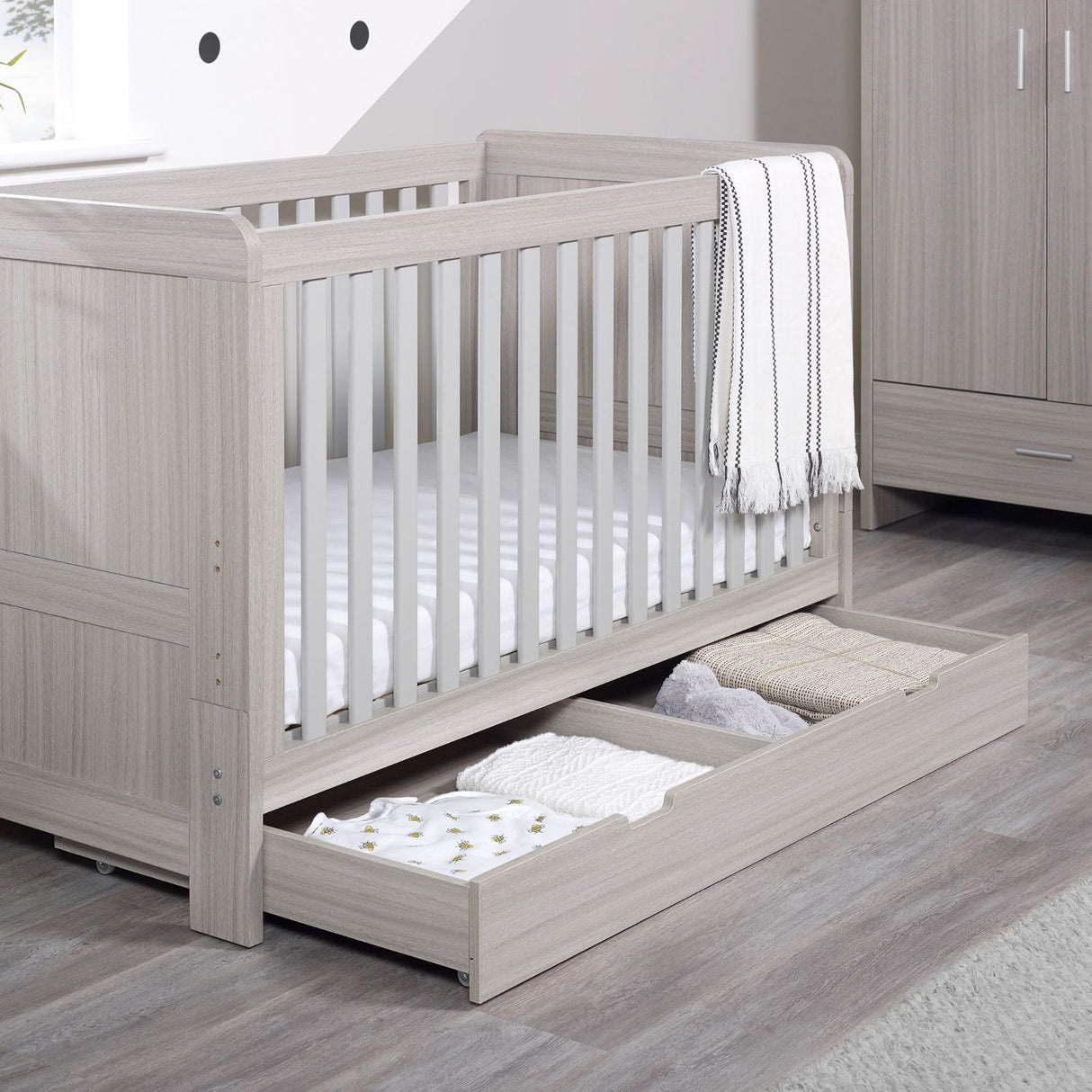 Angled lifestyle shot of Ickle Bubba Pembrey Cot Bed and All Seasons Premium Pocket Sprung Mattress set with opened Under Drawer storage full of clothes in Ash Grey colour