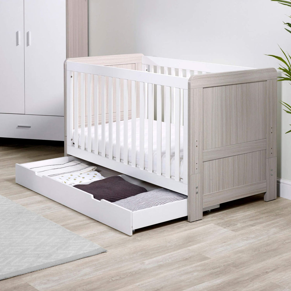 Angled lifestyle shot of Ickle Bubba Pembrey Cot Bed with opened Under Drawer storage full of clothes in Ash Grey and White colour