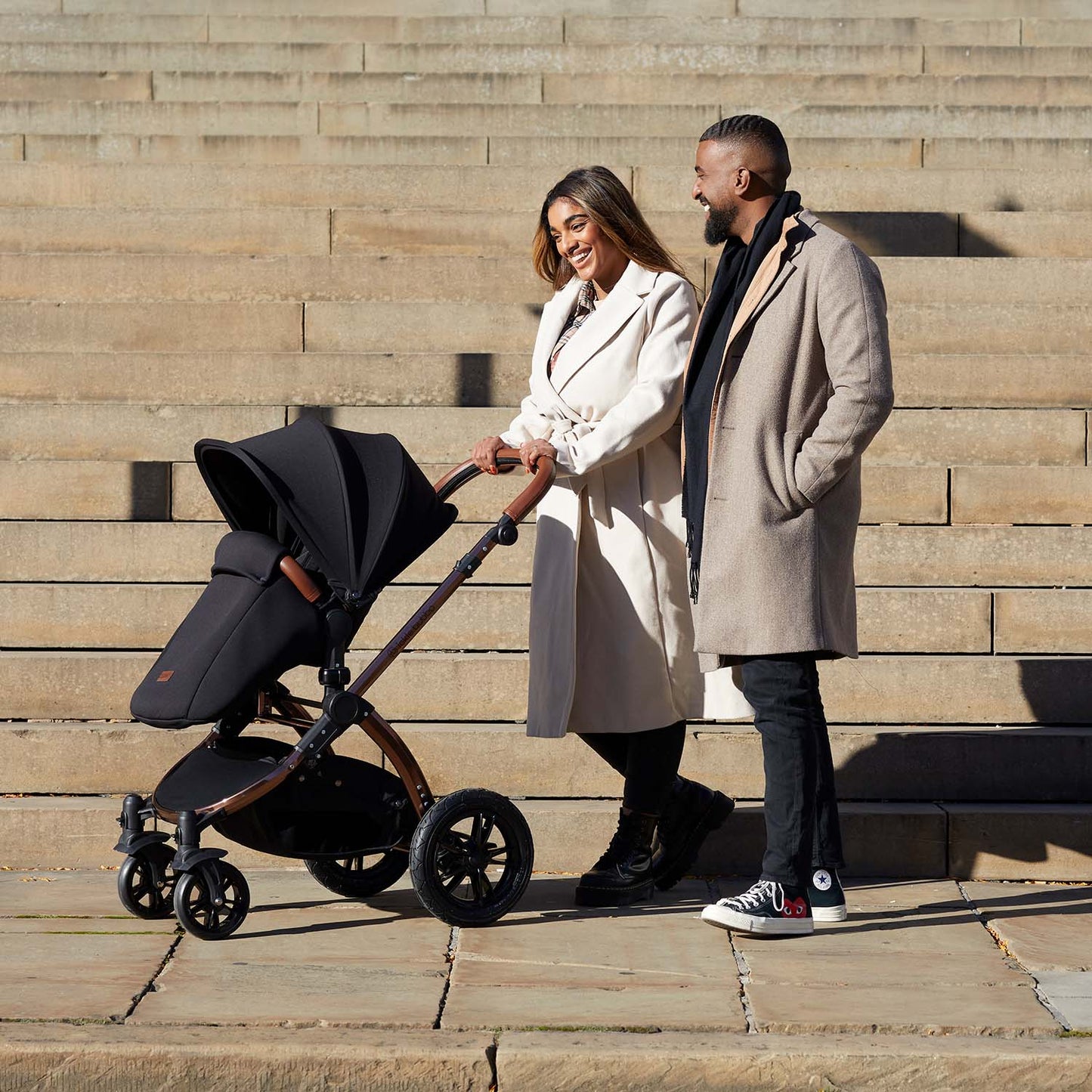 A couple walking while pushing an Ickle Bubba Stomp Luxe All-in-One Travel System in Midnight black colour