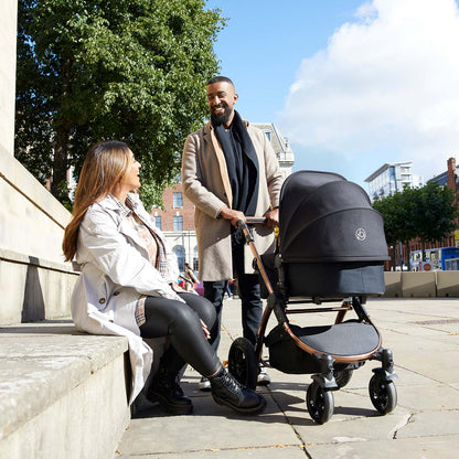 A smiling man holding an Ickle Bubba Stomp Luxe Pushchair with carrycot beside a sitting woman