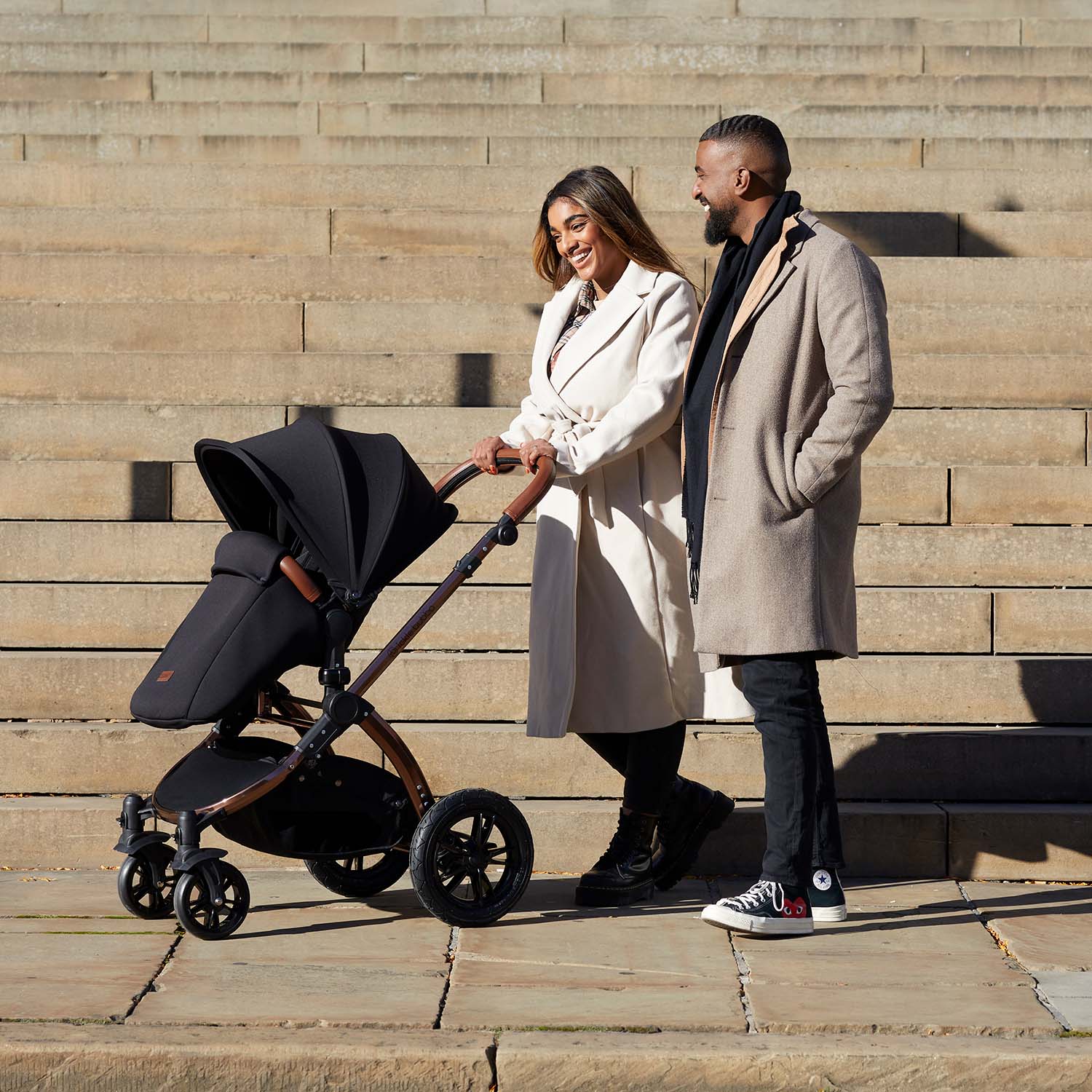 A smiling couple walking while pushing an Ickle Bubba Stomp Luxe All-in-One Travel System in Midnight black colour