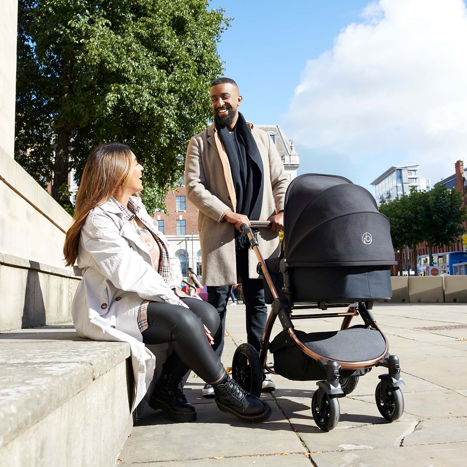 A smiling man holding an Ickle Bubba Stomp Luxe Pushchair with carrycot in Midnight colour beside a sitting woman