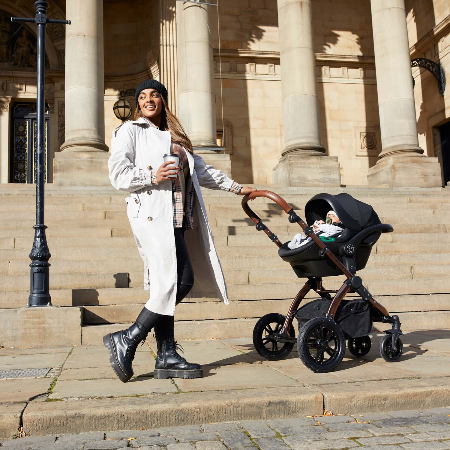 A woman holding a coffee cup while pushing an Ickle Bubba Stomp Luxe All-in-One Travel System in Midnight black colour