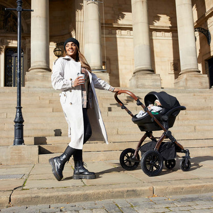 A woman holding a coffee cup while pushing an Ickle Bubba Stomp Luxe Pushchair with Stratus i-Size car seat attached with a baby sleeping