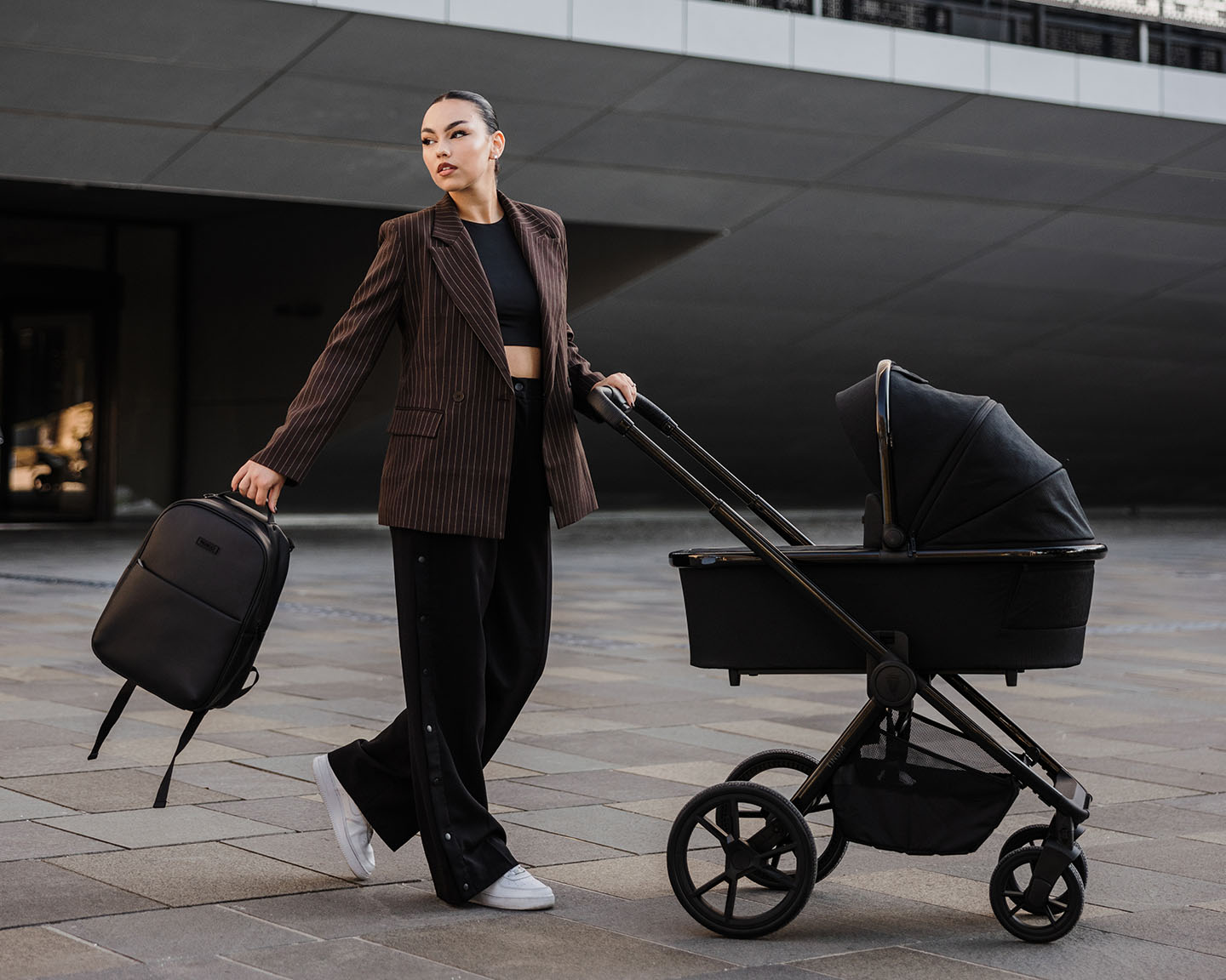 a woman pushing the Venicci Tinum Edge SE Raven in one hand while holding the Backpack in another hand