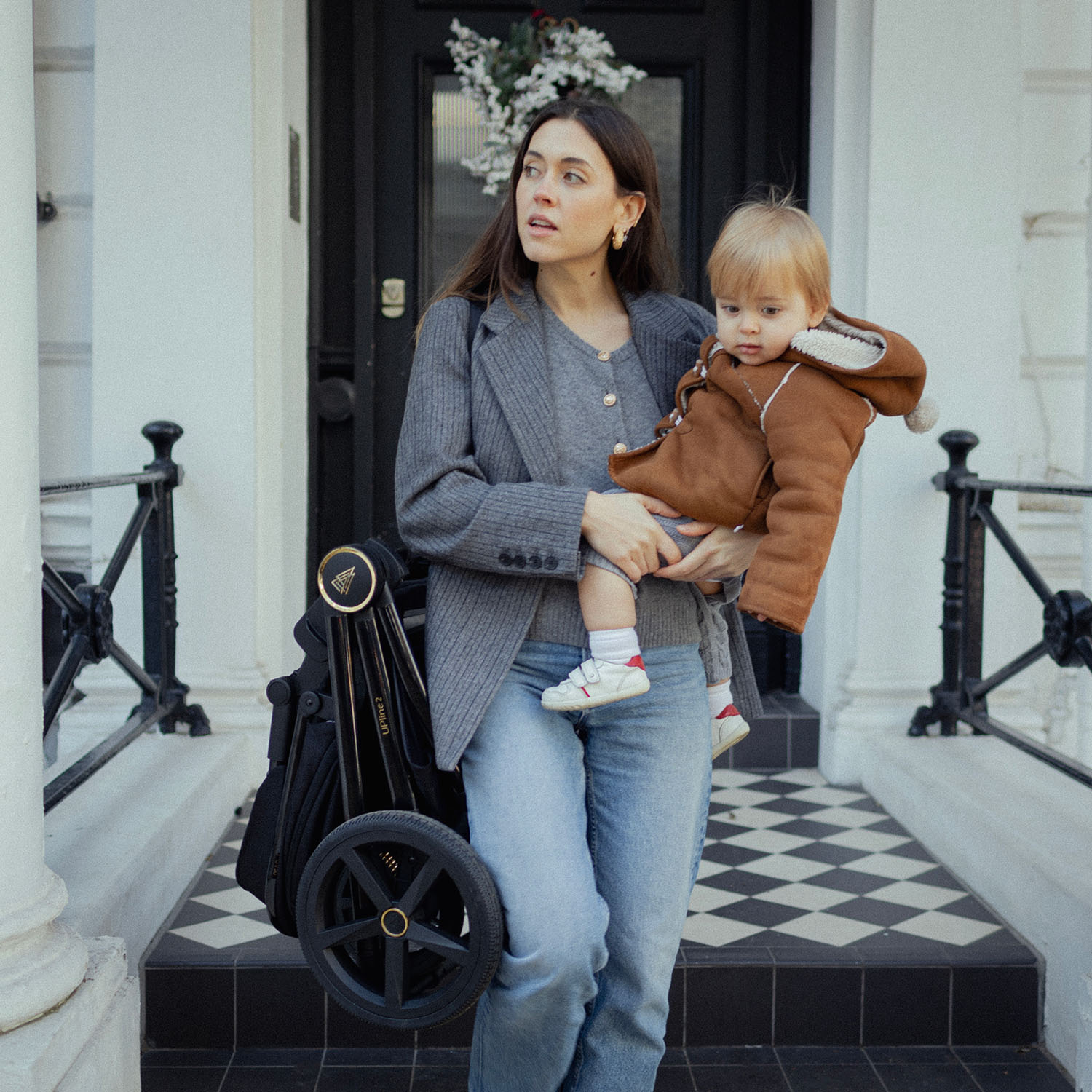 A mother carrying both her baby and a folded Venicci Upline 2 Pushchair in All Black colour